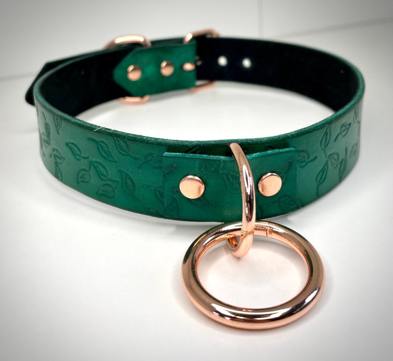 K&ouml;nnte beinhalten: Ein gr&uuml;nes Lederhalsband mit einem gepr&auml;gten Blattmotiv in Ros&eacute;gold und einem daran befestigten Ros&eacute;goldring.