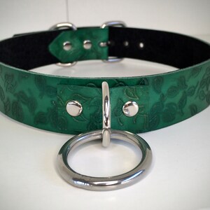 K&ouml;nnte beinhalten: Ein gr&uuml;nes Lederhalsband mit Blumenmuster und einem silbernen Ring. Das Halsband hat ein schwarzes Futter und silberne Beschl&auml;ge.