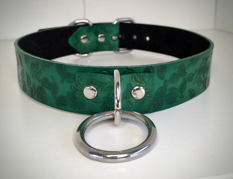 K&ouml;nnte beinhalten: Ein gr&uuml;nes Lederhalsband mit Blumenmuster und einem silbernen Ring. Das Halsband hat ein schwarzes Futter und silberne Beschl&auml;ge.