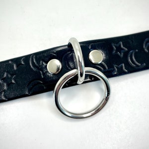 May include: Black leather choker with a silver metal ring and a crescent moon and star pattern.