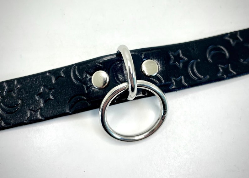 May include: Black leather choker with a silver metal ring and a crescent moon and star pattern.