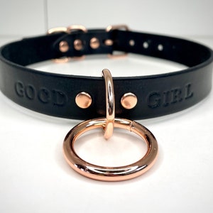 May include: Black leather choker with rose gold hardware and the words "GOOD GIRL" embossed on the front.