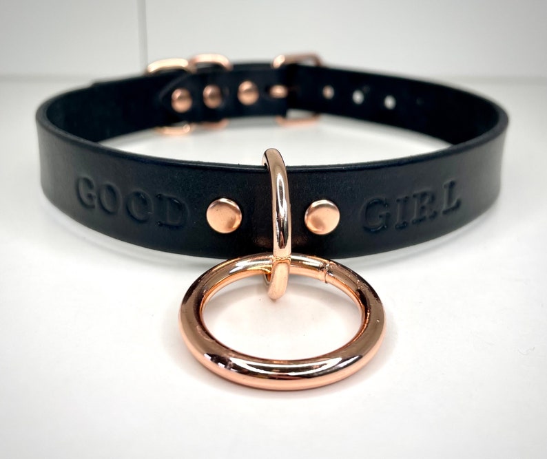 May include: Black leather choker with rose gold hardware and the words "GOOD GIRL" embossed on the front.