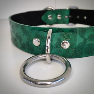 K&ouml;nnte beinhalten: Ein gr&uuml;nes Lederhalsband mit einem gepr&auml;gten Blumenmuster und einem silbernen Metallring. Das Halsband hat ein schwarzes Futter und silberne Metallbeschl&auml;ge.