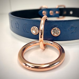 May include: A navy blue leather choker with rose gold hardware and a decorative ring. The choker has a star and crescent moon design and clear rhinestones.