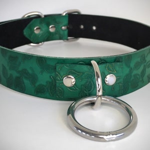 K&ouml;nnte beinhalten: Ein gr&uuml;nes Lederhalsband mit einer silbernen Schnalle und einem gro&szlig;en silbernen Ring. Das Leder ist mit einem Blumenmuster gepr&auml;gt.