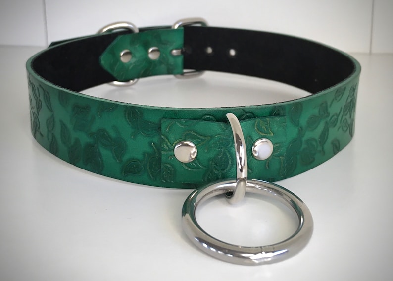 K&ouml;nnte beinhalten: Ein gr&uuml;nes Lederhalsband mit einer silbernen Schnalle und einem gro&szlig;en silbernen Ring. Das Leder ist mit einem Blumenmuster gepr&auml;gt.