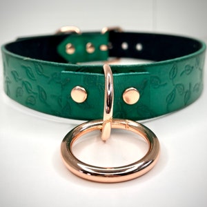 K&ouml;nnte beinhalten: Ein gr&uuml;nes Lederhalsband mit einem ros&eacute;goldenen Metallring und einem Blattmotiv.