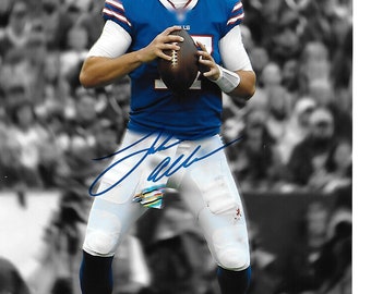 May include: A football player wearing a blue jersey with red and white sleeve stripes, white pants, and a silver helmet, holding a brown football. The image features a blue ink autograph across the center and a blurred stadium background.