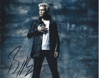 May include: A studio portrait of a person with spiky, bleached blonde hair, wearing a black leather jacket over a white t-shirt and dark jeans. The person is standing with one hand on their chest. The image is signed with a black marker.