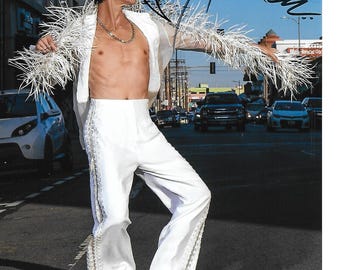 May include: A person wearing a white feathered jacket and white pants with a white trim. They are wearing red high heels and have a gold chain necklace around their neck. The person is posing in a street with cars in the background.