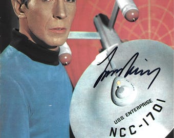 May include: A photograph of a character with pointed ears wearing a blue long-sleeve shirt with a black collar, holding a model of the U.S.S. Enterprise NCC-1701 starship. The model features a black ink signature across the saucer section.