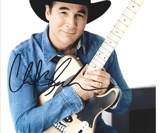 Könnte beinhalten: Ein Porträt eines Mannes mit schwarzem Cowboyhut und Jeanshemd, der eine hellfarbene E-Gitarre hält. Die Gitarre hat ein schwarzes Schlagbrett und silberne Hardware. Das Bild ist mit einem schwarzen Marker signiert.