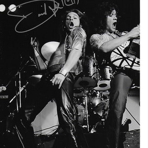 May include: Black and white photo of two musicians performing on stage. The musician on the left is singing and the musician on the right is playing a guitar with a black and white striped design. The signature "Eric Bell" is written in the top left corner of the photo.