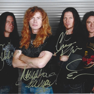 May include: A photograph of four individuals standing in a row, each wearing a black t-shirt. The person second from the left has long, wavy red hair. Each individual has a signature written in gold ink across their chest area on the photograph.