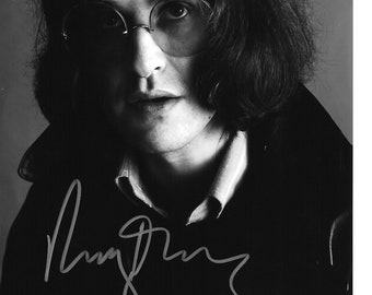May include: Black and white portrait of a person with long hair, round glasses, and a dark coat. The image includes a signature in the lower left corner. The person is looking directly at the camera.