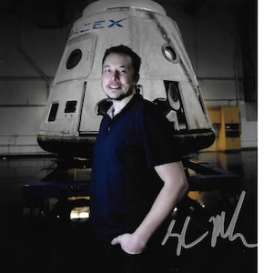 May include: A photograph featuring a man in a dark blue polo shirt posing in front of a spacecraft. The spacecraft is white with the text "SPACEX" visible. The man is smiling and the image includes an autograph.