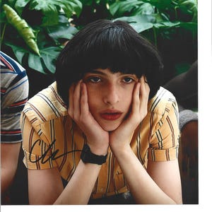 Peut inclure: Portrait d'une personne aux cheveux foncés portant une chemise jaune à rayures, posant son menton sur ses mains. La chemise a des manches retroussées et une signature est visible. Un feuillage vert luxuriant forme l'arrière-plan.