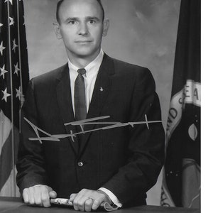 May include: Black and white portrait of a man in a suit and tie, standing in front of two flags. He is holding a rolled-up document in his hands. The man is smiling and looking at the camera. The flags are the American flag and the NASA flag. The man's signature is visible in the lower left corner of the image.