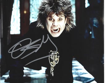 May include: A glossy photograph featuring a person with shoulder-length, layered brown hair, wearing a black long-sleeved top with a decorative gold-toned pendant necklace. The person has an open-mouthed expression and a silver signature is visible.
