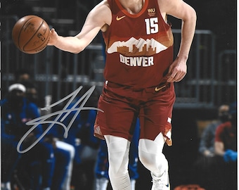 May include: A basketball player wearing a maroon Denver jersey with number 15 and a mountain graphic, white compression leggings, and white sneakers, dribbling a brown basketball. The image features a prominent black ink autograph across the front.