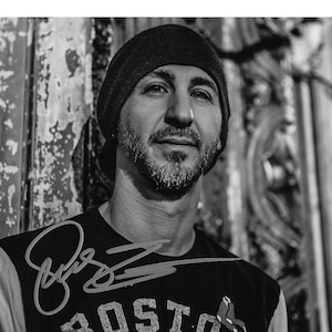 Puede incluir: Foto en blanco y negro de un hombre con una boina negra y una camiseta negra con la palabra "BOSTON" impresa en ella. El hombre mira a la cámara y tiene barba. La camiseta tiene una firma.