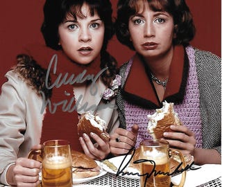 May include: A vintage photograph featuring two women holding partially eaten burgers and mugs of beer. They are seated at a table with food and autographs. One woman wears a red scarf and the other a purple sweater.