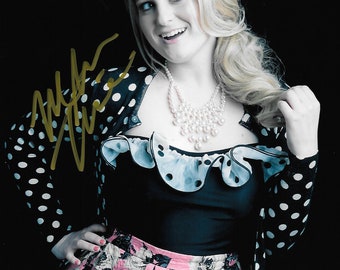 May include: A woman with blonde hair wearing a black top with a ruffled collar and a pink floral skirt. She is also wearing a black polka dot cardigan and a pearl necklace. The image has a black background and a signature in gold.