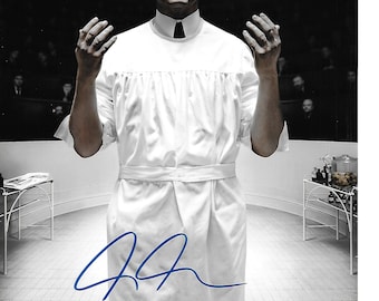 May include: A black and white photo of a man in a white robe with his hands raised. The man has a mustache and is looking at the camera. The photo is signed with a blue signature.