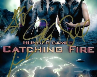 May include: A movie poster for the film "The Hunger Games: Catching Fire" featuring Jennifer Lawrence, Josh Hutcherson, and Liam Hemsworth. The poster has a blue and green color scheme with a large image of a glowing sphere in the center. The title of the film is written in large, white letters at the bottom of the poster.
