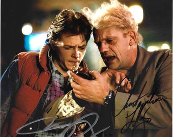 May include: A glossy photograph featuring two individuals looking at a small round clock. One person wears a red puffer vest over a plaid shirt, while the other has wild white hair and a tan jacket. The image includes two distinct black ink signatures.