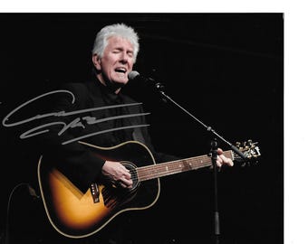 May include: A man with white hair sings into a microphone while playing an acoustic guitar. The guitar has a sunburst finish and is signed. The man is wearing a black suit and is on a stage with a dark background.