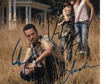 May include: A signed photograph of three actors from the television show The Walking Dead. The actors are Andrew Lincoln, Chandler Riggs, and Lauren Cohan. The photo is taken outdoors in a field with a house in the background.