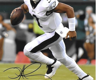 May include: A football player wearing a white jersey with a black stripe down the side of each leg, a black and white helmet, and white shoes runs with the football in his hands. The player is wearing a white wristband on his left wrist. The player is on a green field with a crowd of people in the background.