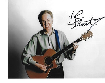 May include: A man smiles while holding an acoustic guitar and playing it. He is wearing a light brown shirt and a blue strap. The guitar is brown with a black pickguard. The man's signature is in the top right corner of the image: "Al Stewart".