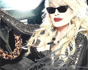 May include: A signed photograph of a blonde individual with voluminous hair wearing black cat-eye sunglasses, a black lace-trimmed top, and black gloves. The individual is seated in a vehicle with a leopard print steering wheel cover and a signature.