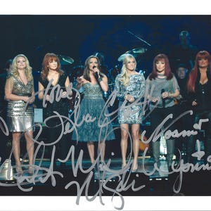 May include: Seven women stand on a stage, each holding a microphone. They are dressed in stage outfits, including dresses and tops. The image is signed with multiple signatures. The background includes stage lighting and musical instruments.
