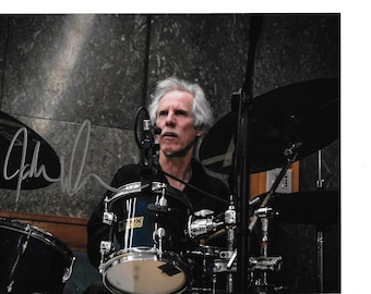May include: A black and white photo of a man playing drums. He is wearing a black shirt and has gray hair. The drums are in focus and the man is slightly out of focus. The photo is signed "John" in the bottom left corner.