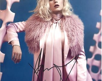 May include: A portrait of a person with blonde hair wearing a pink outfit, including a blazer, pants, and a top with a necktie. The outfit is accented by a fluffy pink collar. The background is blue with pink geometric shapes and an autograph.