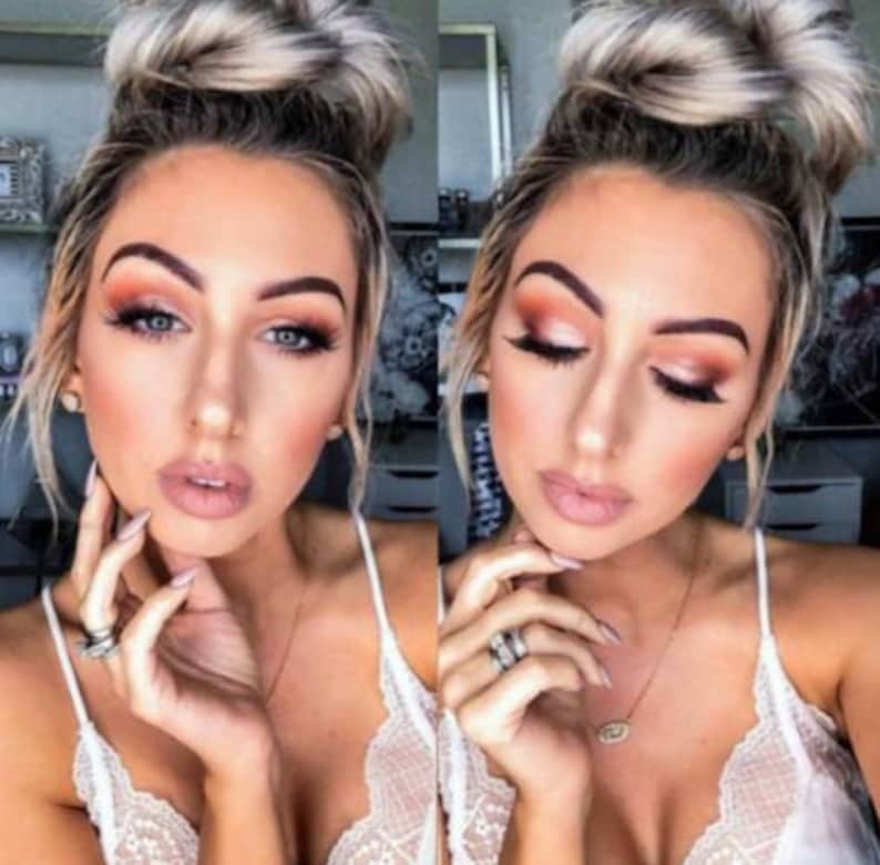 May include: A woman with light brown hair styled in a bun, wearing a white lace top, has on natural makeup with a peachy eyeshadow and a nude lip color. She is wearing a gold necklace with a small oval pendant.