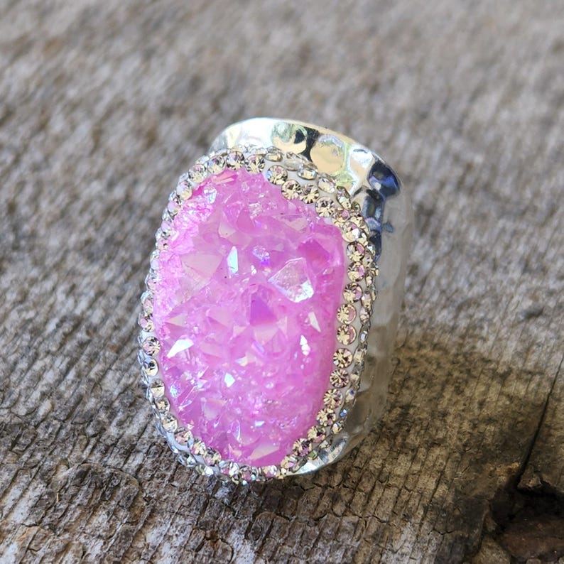 May include: A silver ring with a large pink crystal set in a clear resin base. The crystal is surrounded by a row of small clear rhinestones.