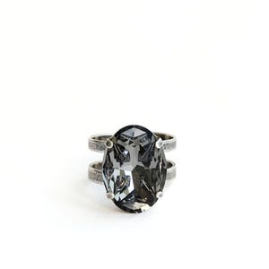 May include: A ring featuring a large, oval-shaped, faceted dark gray crystal set in a prong setting. The band is constructed from two parallel, textured silver-toned metal strips that connect to the central stone setting on both sides.