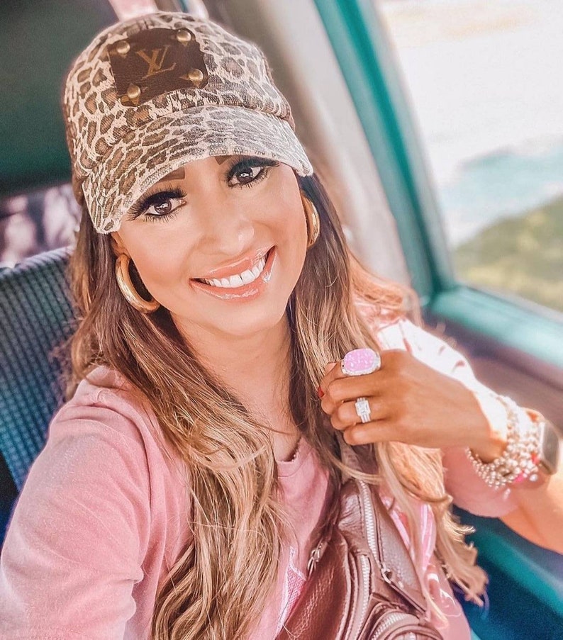 May include: A woman wearing a pink shirt and a leopard print baseball cap with a brown leather patch that says "LV". She is wearing gold hoop earrings and a pink ring with a large stone.