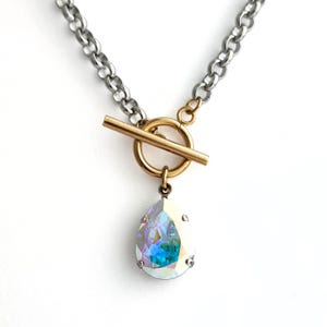 Peut inclure: Un collier à chaîne argenté avec un fermoir à bascule doré et un pendentif en cristal irisé en forme de poire. Le pendentif reflète des nuances de bleu, vert et violet. Sur fond blanc.