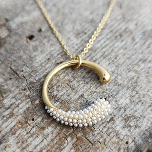 May include: A gold necklace with a curved pendant. The pendant is made of a gold circle with a row of small white pearls around the bottom half.