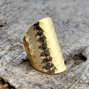 May include: A gold hammered metal ring with a wide band. The ring is curved and has a textured surface.