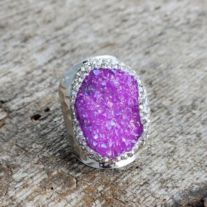 May include: A silver ring with a large, oval-shaped purple gemstone surrounded by clear rhinestones.