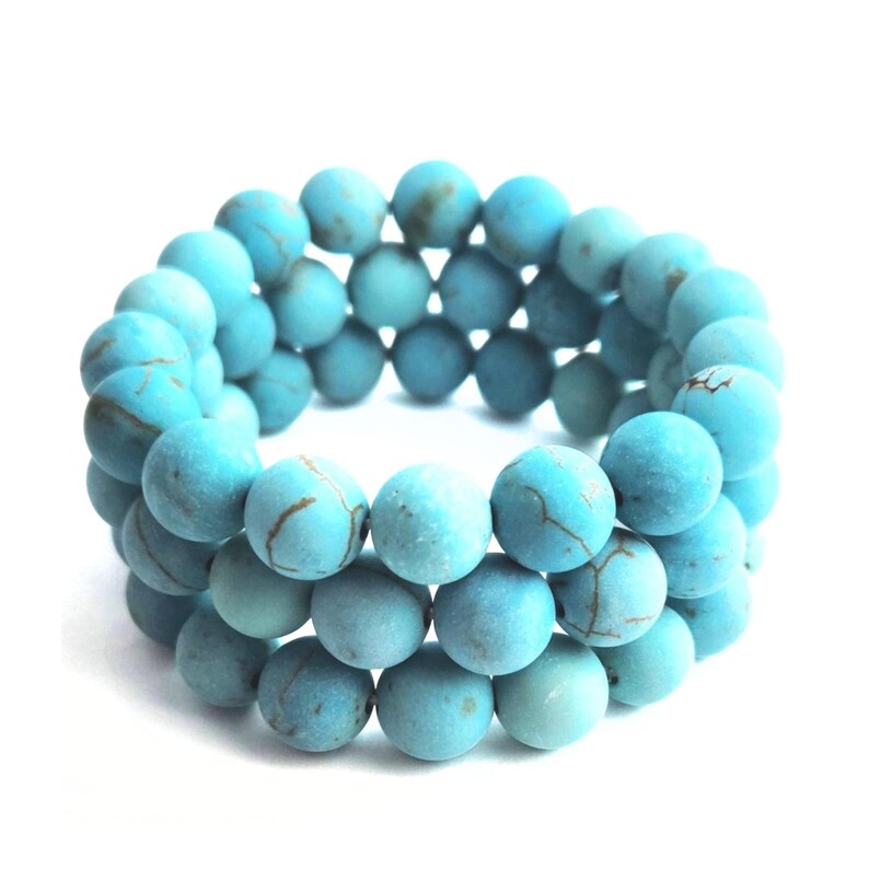 Puede incluir: Una pulsera de tres hilos compuesta por cuentas redondas de color turquesa con acabado mate. Cada cuenta presenta patrones de vetas marrones de aspecto natural en toda la superficie, creando una textura consistente en las tres filas circulares.
