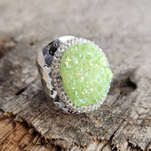 May include: A silver ring with a large, green, iridescent gemstone surrounded by a row of clear rhinestones.