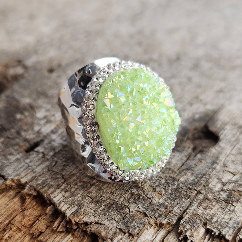 May include: A silver ring with a large, green, iridescent gemstone surrounded by a row of clear rhinestones.